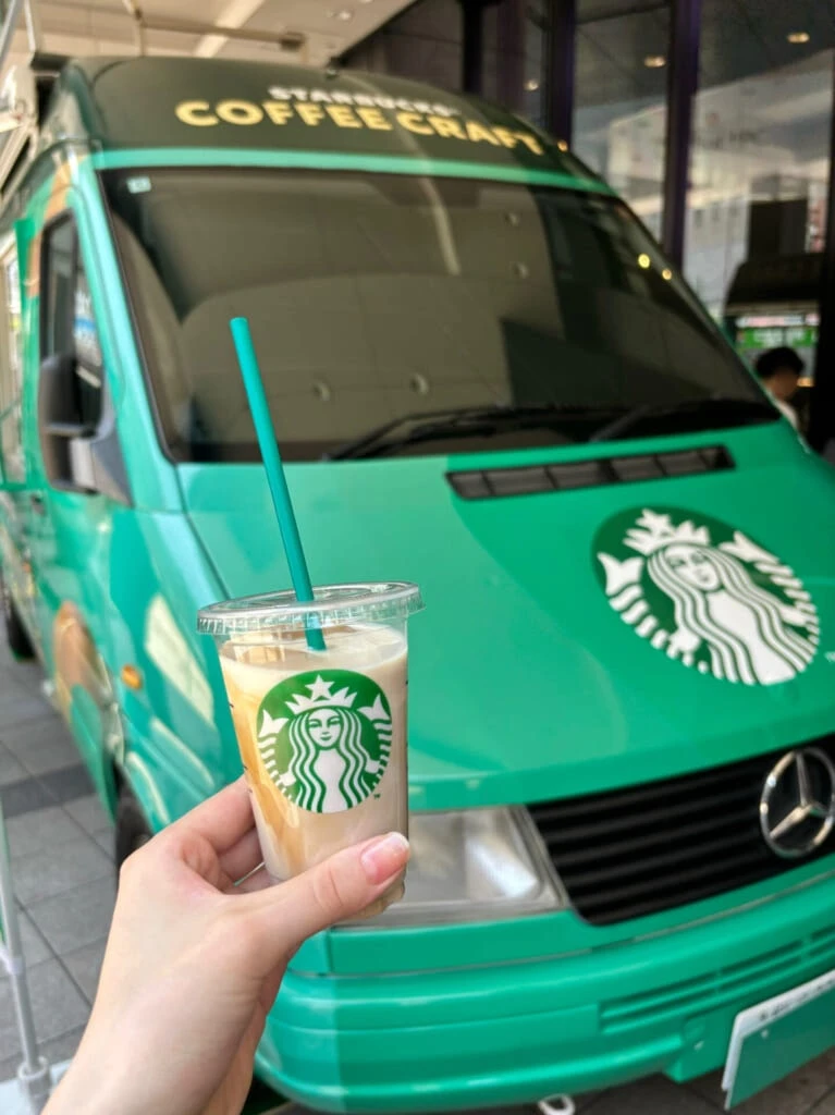 スタバグリーンのキッチンカーを背景にキャラメルマキアートを飲む