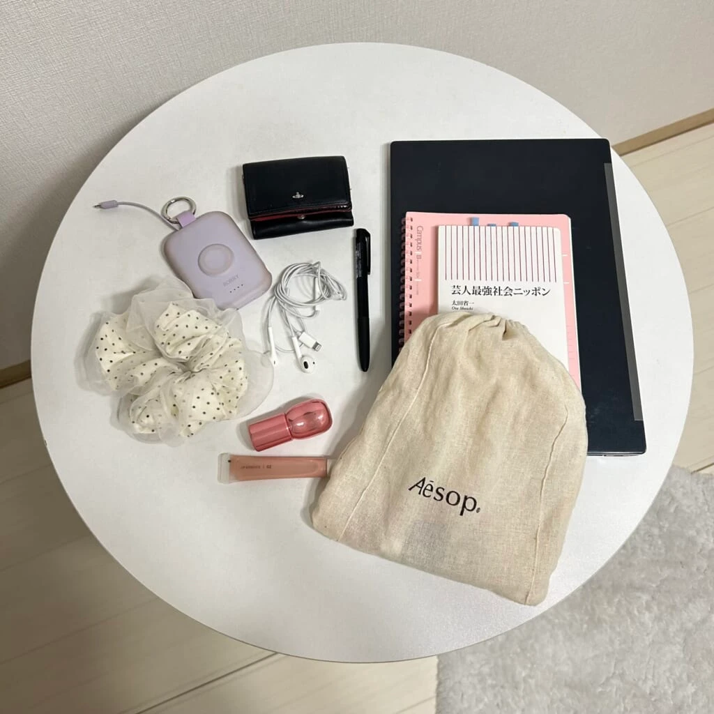 シュシュ、モバイルバッテリー、有線のイヤホン、財布、黒のボールペン、パソコン、ノート、本、ポーチ、保湿リップ、グロスを並べて上から撮影した写真。